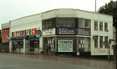 A corner building with the exterior painted white, housing a number of shops: ½ Price Jewellery, Betterspecs, City Centre Bureau, and Fingertips Nail Care Studios. Fingertips is on the first floor above City Centre Bureau. The sky is grey and the road looks wet with rain. There are three pedestrians, two of them sharing an un=mbrella.