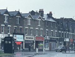 A terrace of three-storey brick buildings with pointed gables. The ground floors are all shops — Michigan Cleaners, Broomfields, United Dairies, Meyers, and others that are too far away from the camera to make out. A black police box is visible to the left.