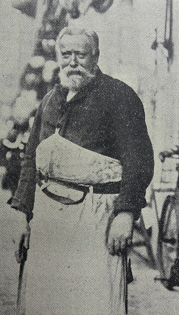 A black-and-white photo of a sturdily-built man with white hair and beard, wearing dark overalls and a white apron.