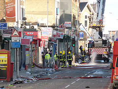 A view along a road with a fire engine and a number of firefighters standing around in the middle distance.  Shop signs cluster along the left side of the photo, including one reading “Kerr”.  Debris and litter lie along the road.