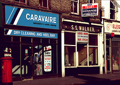 Two terraced shopfronts, both with large front windows and signs above.  The left-hand shop has a light blue sign reading “Caravaire / specialists in curtain cleaning / DRY CLEANING AND HEEL BAR” above the frontage and a poster in the window reading “2 hr dry cleaning / Saturdays included / plus first class shoe repairs”.  The right-hand shop has a rather more old-fashioned sign reading “S. S. Walker.” above the frontage and another sign above the central door reading “Ladies & mens / S S Walker / Hair stylist”.  The right-hand shop also has a projecting estate agent’s sign higher up, reading “Shop to let / Stuart Edwards & Partners / 102/4 High St Croydon / 01688 8313”.  A small slice of another shop can just be seen at right of frame, with the number “34” and the words “ELEG... / Ladies & G...”.