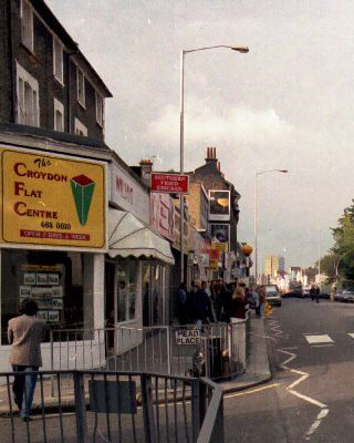 A view along a terrace of shops, taken end-on from the corner opposite.  The shop in the foreground has a diagonal corner with a sign reading “The Croydon Flat Centre”.