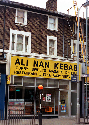 A terraced shop with a large black-on-yellow sign above reading “Ali Nan Kebab / Curry Sweets Masala Dhosai / Restaurant & Take Away Serv[...].  Orange plastic chairs are visible through the fully glazed shopfront.”