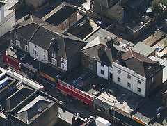 An oblique aerial view of shops along one side of a road.  All of them have been extended forwards as far as the road on the ground floor, though some of the buildings (including number 59) were built further back than others, so their extensions are correspondingly longer.