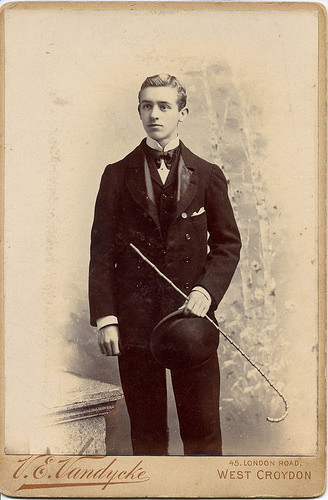 A sepia photo of a young, white-looking man with slicked-back hair.  He’s dressed in a suit with a high collar and bow-tie, and he’s holding a cane and a bowler hat.  Printed at the bottom of the mount is the name “V. E. Vandycke” in cursive, and the address “45, London Road, West Croydon” in a plainer font.