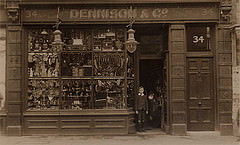 A sepia-toned photograph showing a single terraced shopfront with “Dennison & Co” on the sign above and a large number 34 on the side door.  A young boy stands in the doorway of the shop.  The shop windows are crammed full of stock.