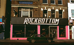 A terraced shop stretching over two shopfronts.  The frontage is painted pink, with a large white-on-black sign above reading “Rockbottom” in capital letters.  Visible in the windows are acoustic guitars, drums, and signs reading “Tonebanks” and “Postex”.