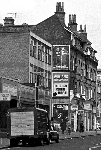 A black-and-white photo with an oblique view onto a 1960s office block next to a 1890s four-storey terrace.  Three advertisement posters are visible on the side wall of the terrace.