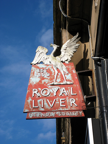 A painted metal projecting sign reading “Est 1850 / Royal Liver Friendly Society”.  Much of the (red) paint has worn off. A stylised “Liver bird” (a cormorant with a frond in its mouth) forms the top of the sign.