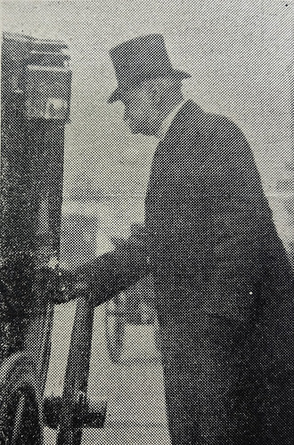A black-and-white photo of a white man in a top hat and dark coat, seen sideways on, with his hand on the door of a carriage.  He appears not to have noticed the photographer.