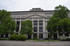 A large grey building with decorative multi-storey pillars, partially obscured by trees.