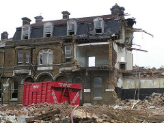 A partly-demolished building with rubble and a large red skip in the foreground.