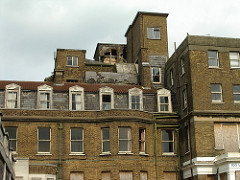 The upper floors of a brick building.  Some of the windows have glass missing, others are boarded up, and the roof is in disrepair.