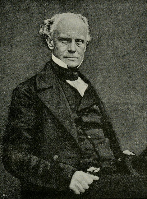 A black-and-white photograph of a white man in his 50s or 60s with receding white hair and a neutral expression.  He is seated and wearing a dark suit.