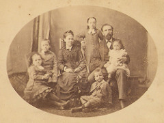 An oval-shaped sepia photograph showing a woman, a man, and five children, all white.  All are seated except for the oldest child, who is standing up behind and between the adults and leaning on the back of the woman’s chair.  The man has a moustache and full beard, and has the youngest child sitting on his lap.  The woman has a slight smile, while the others have fairly fixed expressions.