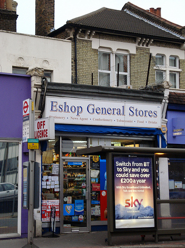 The same terraced shop as in the first picture in this article, but taken from a different angle and with a different sign; this one reads “Eshop General Stores”, with “Stationery · News Agent · Confectionery · Tobacconist · Food · Drinks” underneath, and is on a white background.  A projecting sign on the left of the frontage reads “OFF LICENCE”.