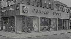 A black-and-white photo of a car showroom located on the corner of two streets and stretching over three shopfronts.  Large windows reveal cars parked inside.  The name “Donald Vince” is in large capital letters along two sides of the showroom, and Volswagen, Volvo, and Daimler logos are on the corners of the signs.
