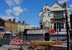 Part of a terrace of shops in the process of being demolished.  The shop on the left, which has a sign reading “G T Stewart / Solicitors & Advocates”, has been reduced to a single storey.  The shop next to it retains at least the facade of its two upper storeys, showing arch and balcony detailing and a peaked roof.  Barriers block off the road in front.