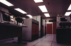 A room full of computer equipment, with large rectangular ceiling lights and utilitarian tiling on the floor.  No windows are visible.  The computers themselves are large, cabinet-sized and -shaped, in white and orange.