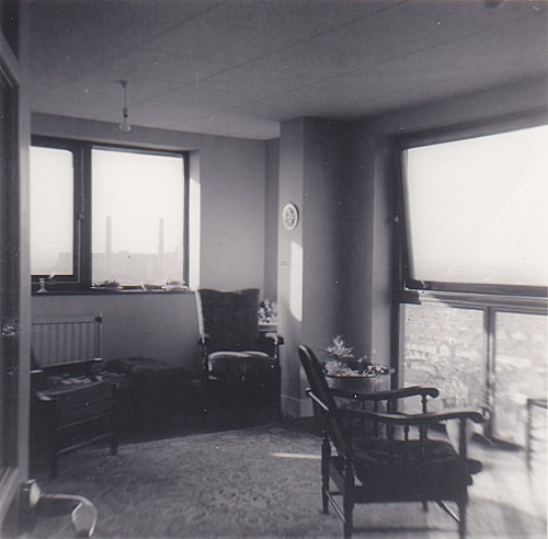A small living room with large windows on two sides showing views consisting mainly of sky.  A couple of wooden armchairs sit on a patterned rug, and a bare bulb hangs from the ceiling.