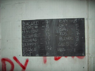 A dirty white wall with parts of spraypainted red words just visible.  A blackboard shows chalked names of drinks along with numbers such as “Bacardi — 7”, “Scotch — 11”, “Vodka — 15½”, and “Gin — 6”.