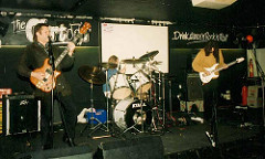 Three white people on stage playing bass guitar, drums, and guitar.  The bass player is singing into a microphone.  Behind the band, the phrases “The Cartoon” and “Drink, dance ‘n’ Rock ‘n’ Roll” can be seen on the wall.  The audience are not visible.