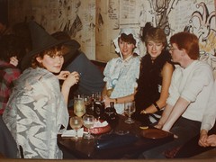 Four light-skinned people sitting around a table, three of them in fancy dress.  Full and empty glasses are clustered around an ashtray on the table.  The white wall behind them is covered with hand-drawn cariacatures and cartoons.