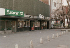 The other end of the long, two-storey concrete block shown in the first image in this article.  The three shopfronts at the right-hand side have been amalgamated into one, with a sign reading “Cartoon” across the middle.  There are two sets of double entrance doors, with windows between.  To the left of this is a shuttered shopfront with a “Europcar” sign above.