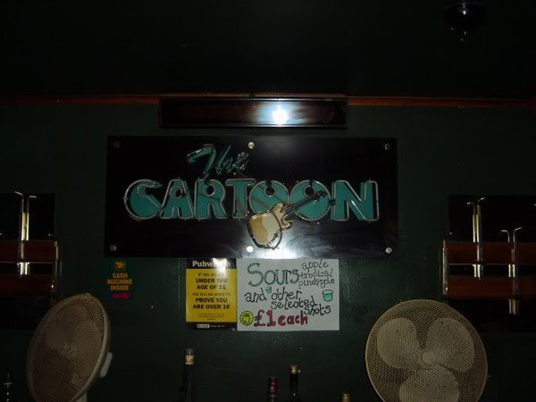 A dark-painted interior wall with “The Cartoon” painted on it and a sign offering “Sours and other selected shots” at “£1 each”.  A couple of large electric fans can be seen in the foreground.