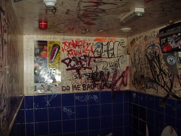 Three walls of a small room with blue tiling below and light-painted walls above.  The walls are covered with graffiti.  The edge of a urinal can just be seen at the bottom of the image.