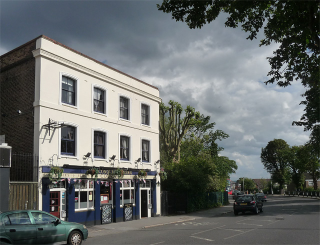 A square three-storey building with the upper floors painted white and the ground floor in dark blue with hanging baskets, blackboards covered with chalk writing, bunting, and the name “Conquering Hero”.  Tall pollarded trees are to the right of the building, and a couple of modern cars are parked outside.  The sky is grey and overcast.