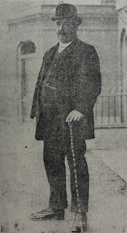 A black-and-white photo of a stocky light-skinned person with a substantial moustache, leaning lightly on a walking cane and wearing a bowler hat and a dark suit with waistcoat and watch chain.