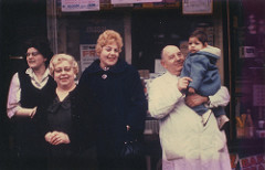 Four adults standing outside a shop window with various notices and boxes of products inside.  One of the adults is wearing a white butcher’s coat and holding a small child.