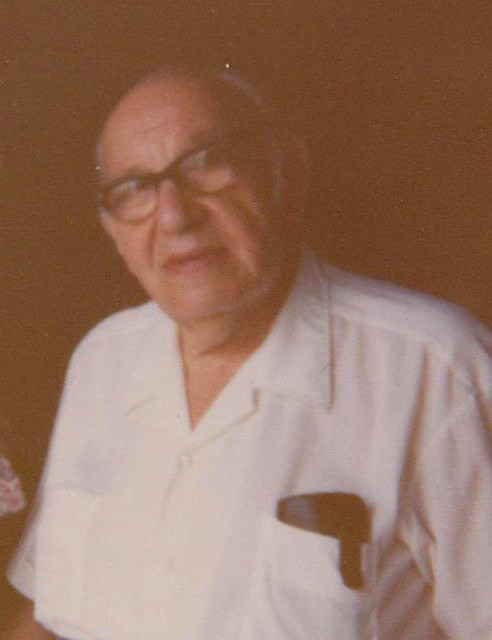 Colour photo of a man wearing black-framed glasses and an open-necked white shirt, looking at the camera with his head slightly turned.