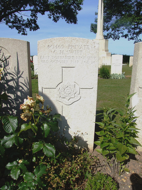 A pale, narrow headstone among others in a grassy graveyard. Decorative shrubs are planted in the foreground.  The engraving reads: “40469 Private C. H. Swift / East Yorkshire Regiment / 3rd October 1917 Age 26”.