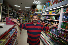 A brown-skinned man with moustache and neatly-trimmed beard, standing in a small shop with his arms held out to the sides.  To the right is a fridge filled with cans and bottles of alcoholic and soft drinks, to the left is a shop counter with a display of chocolate bars in front, and further back are shelves with bottles of wine and spirits.