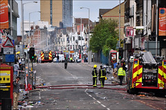 A view northwards along London Road.  The street is strewn with debris and two fire engines are visible, one parked up in the foreground and the other further along with firefighters spraying water on some burned-looking buildings.