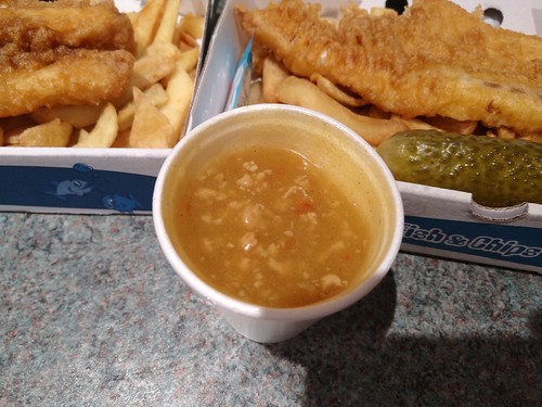 A white polystyrene cup filled with a lumpy light brown sauce.  The corners of two cardboard boxes filled with fish and chips are in the background, one with a gherkin too.