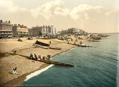A colour postcard showing a shingle beach with groynes stretching into the sea.  A few people are sitting on the beach and the groynes, and several small boats are drawn up on the beach.  A line of four-storey buildings in differing architectural styles can be seen in the background.  Along the bottom is printed “11341.  Worthing.  The beach.  Looking east.”