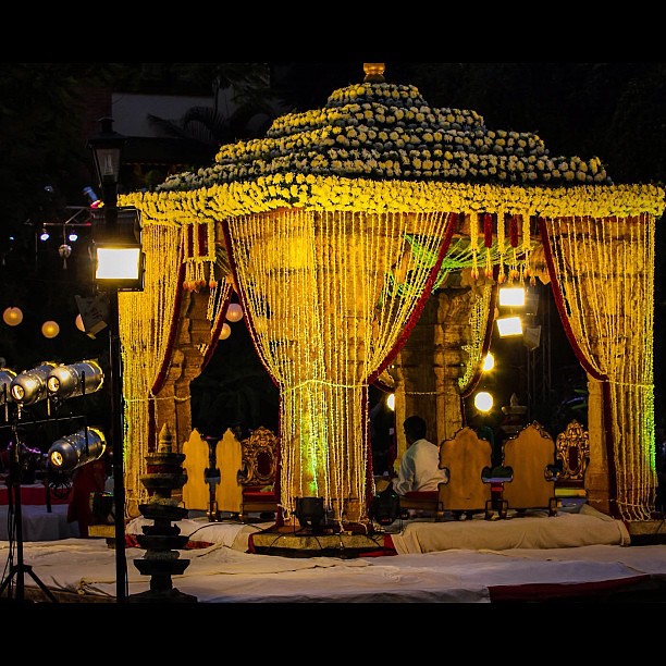 A grand canopy surmounted by tiers of flowers, with long trailing strings of flowers, beads, or similar around the edges, caught back with ties.  Several low seats are underneath, at least one of them occupied. The canopy is on a stage lit by floodlights, while the surroundings are in darkness.