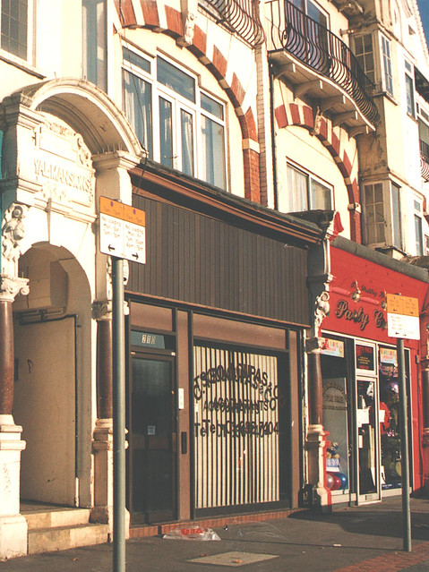 A brown-framed shopfront with decorative arched brickword on the floor above and an arched doorway at the side.  There is no sign above it, but decals on the shop window read “C. Tsouloupas & Co / Accountants”.  Beige window blinds block all view of the interior.