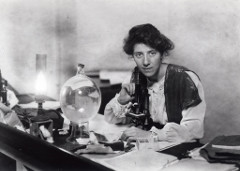 A black-and-white photo of a young white woman seated at a desk with a microscope in front of her.  The desk is covered with pieces of paper and other items of laboratory equipment.
