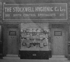 A close-up black-and-white photo of a small ground-floor shop, with doors on either side labelled “Gent’s Entrance” and “Ladies’ Entrance” respectively, and a display window in between crammed full with boxes of various shapes and sizes.  Signs in the window read “Elastic hosiery”, “Trusses”, and “Surgical belts”, while a larger sign above reads “The Stockwell Hygienic Co Ltd / 210 / Birth Control Specialists / 210”.