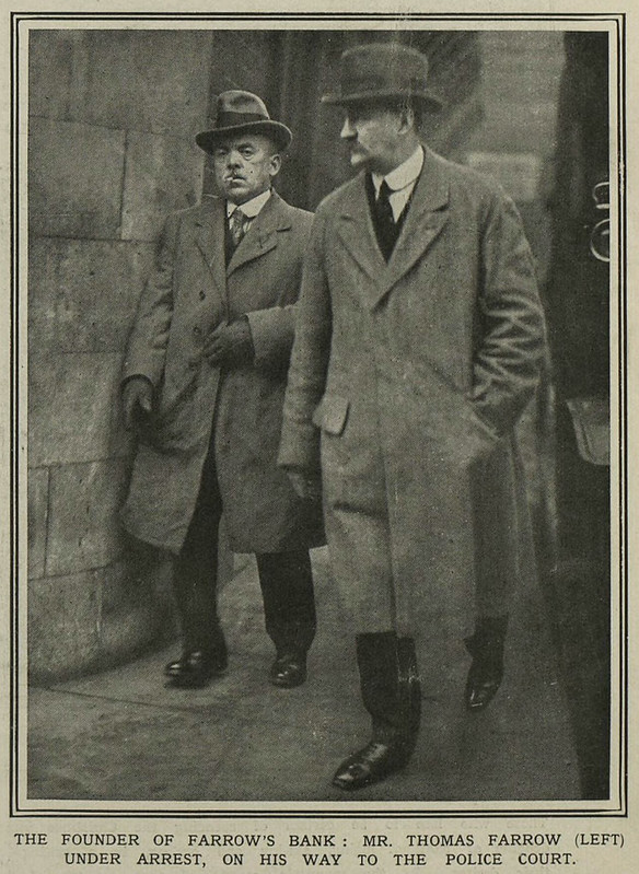 A black-and-white photo of two moustached white men wearing fedoras and long coats over dark trousers, white shirts, ties, and dark shiny shoes.  The man on the left is noticeably shorter than the other, and possibly has a cigarette in his mouth; he is looking at the camera with a slightly grumpy expression.