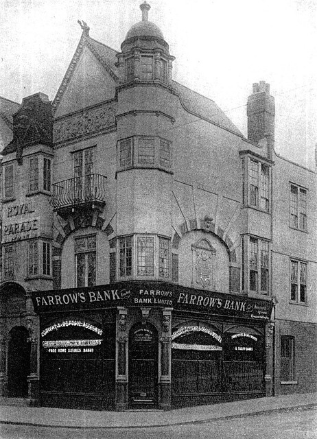 A full-height black-and-white photo of the premises depicted above, showing a dome structure at the very top of the corner profile.  The basic structure of the ground floor is the same, with an arched doorway at the corner and ground-floor windows along both sides, but here the windows have arched inserts at the top and the pillars to either side of the door have additional detailing.  The first-floor window along the side road is absent here, and in its place is an embossed plaque.