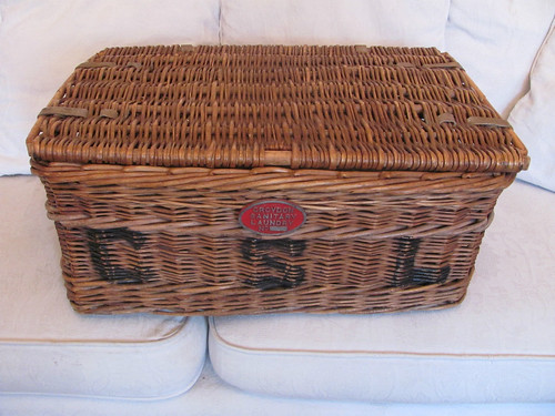 A light brown wicker basket placed on a light-coloured sofa.  The letters “CSL” are painted onto the front of the basket, and a metal plaque above this reads “Croydon Sanitary Laundry”.