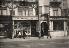 A small terraced shopfront with a sign reading “SportCraft”, styled as overlapping words as in the newspaper advert earlier.  To the right is a recessed archway with the words “Royal Mansions” embossed above, and beyond that is another property with a sign reading “Westminster Bank”.