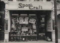 The same shop as earlier, but now the word “SportCraft” on the sign is an outline cut from wood instead of just being painted on.  In the shop window are at least five different posters for the film “Annie Get Your Gun”, along with several toy ride-along horses (and, incongruously, a ride-along snail).  To one side is a recessed doorway with a sign reading “L F Godfrey Coal Order Office”.