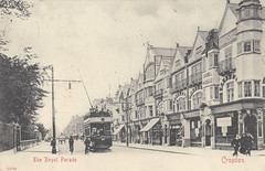 A sepia-toned monochrome photo looking along a road lined on one side with a row of three- and four-story buildings in uniform style.  A brick wall with trees behind runs along the opposite side of the road.  Two or three people are walking along the pavement next to the buildings, and a horse-drawn cart, a trolleybus, and a couple of cyclists can be seen on the road.