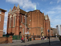 A large, tall red-brick building with long arched windows and an arched entrance.  A copper spire covered with verdigris rises above the roof.  A few people are walking along the pavement outside, making it clear that the building is many times the height of a person.